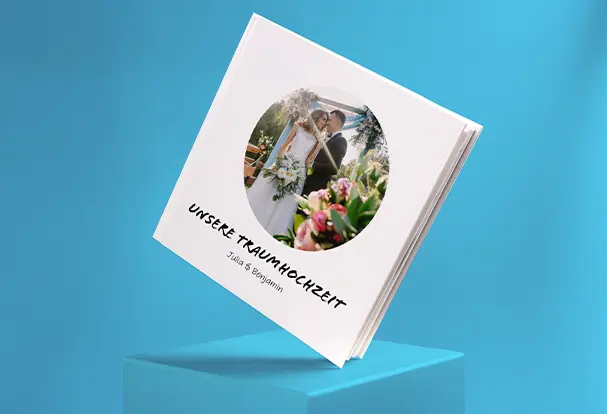 Fotogeschenke Hochzeit: Personalisiertes Fotobuch zur Traumhochzeit Ein quadratisches Fotobuch, auf einer Ecke stehend, mit dem Titel „Unsere Traumhochzeit - Julia und Benjamin“. Dieses individuell gestaltete Fotobuch hält die schönsten Erinnerungen an den besonderen Tag fest und ist das perfekte Fotogeschenk zur Hochzeit.