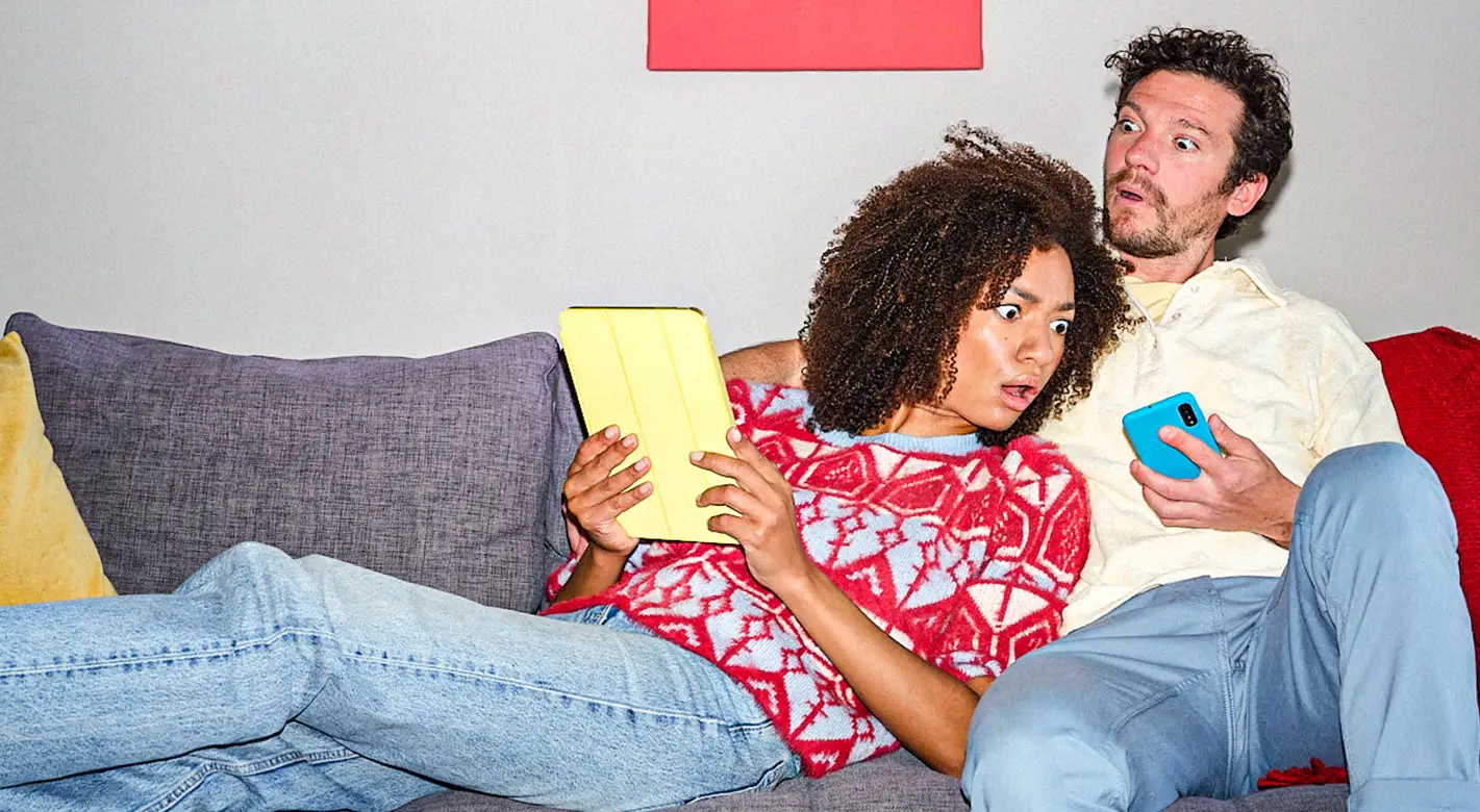 Valentinstag Fotogeschenke – humorvolles Paarmotiv für moderne Liebesgeschenke Valentinstag Fotogeschenke mit humorvollem Paarmotiv: Frau und Mann sitzen auf dem Sofa und schauen überrascht auf Tablet und Smartphone, modernes Alltagsmotiv für persönliche Fotogeschenke.
