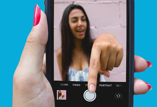 Eine junge Frau greift aus dem Smartphone heraus, um ein Foto von sich selbst zu machen. Unsere kreativen Foto-Tipps zeigen, wie man mit einfachen Mitteln beeindruckende Selbstporträts erzielen kann.
