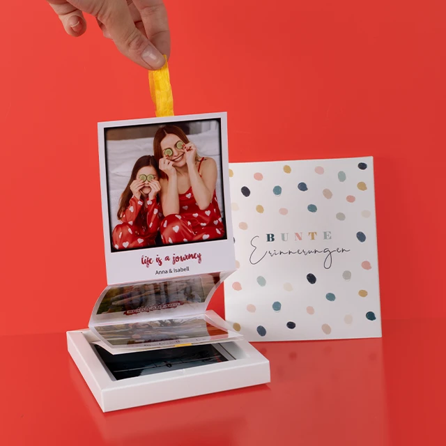 Person zieht Sofortbilder mit gelbem Band aus einer Box, daneben liegt eine farbenfrohe Verpackung mit der Aufschrift „Bunte Erinnerungen“.