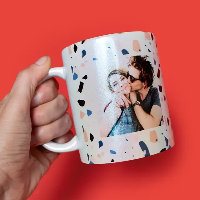 Tasse mit Perlmutteffekt, bedruckt mit Foto Eine Hand hält eine Tasse mit Perlmutteffekt vor einem roten Hintergrund. Die Tasse ist mit dem Foto eines Paares bedruckt.