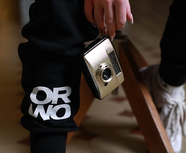 Zu sehen ist, wie jemand eine Retro-Kamera in der Hand hält. Diese Person trägt dabei die stylische Jogginghose von ORWO und repräsentiert so die Retrofotografie-Welt.