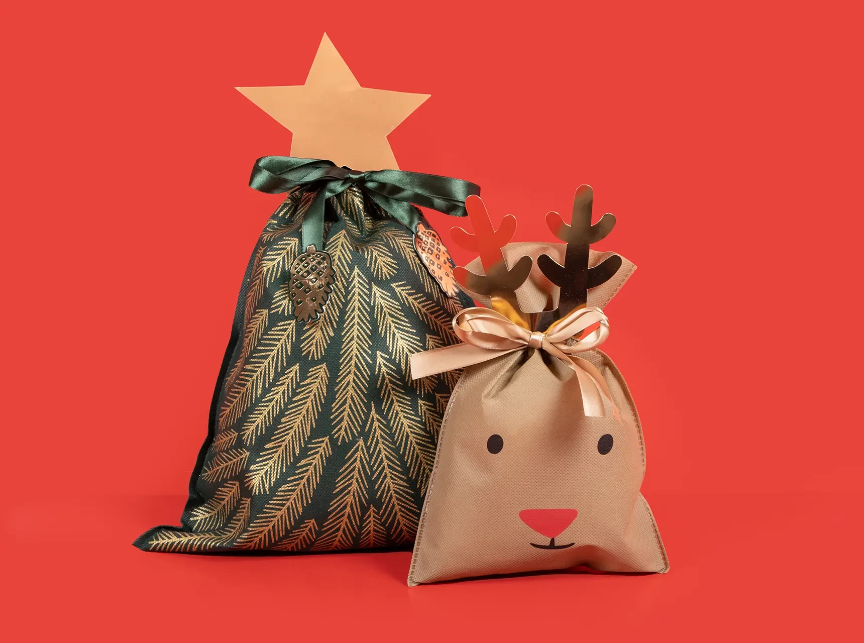 Zwei festliche Geschenktüten: kleines Rentier mit Geweih und große Tüte in Tannenbaumoptik mit Papierstern, beide mit Satinband verschlossen.