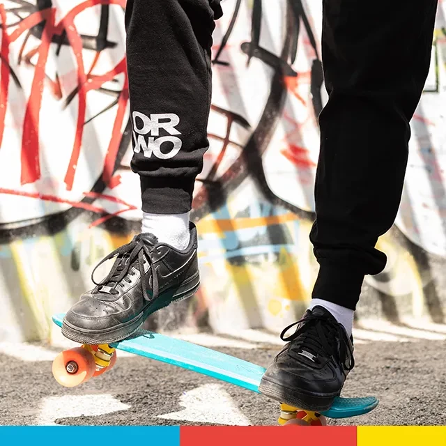 Ein hellblaues Pennyboard ist zu sehen, auf dessen Rücken Beine stehen, die den Jogger von ORWO tragen und diese stylisch in Szene setzen. Im Hintergrund ist Grafitti.