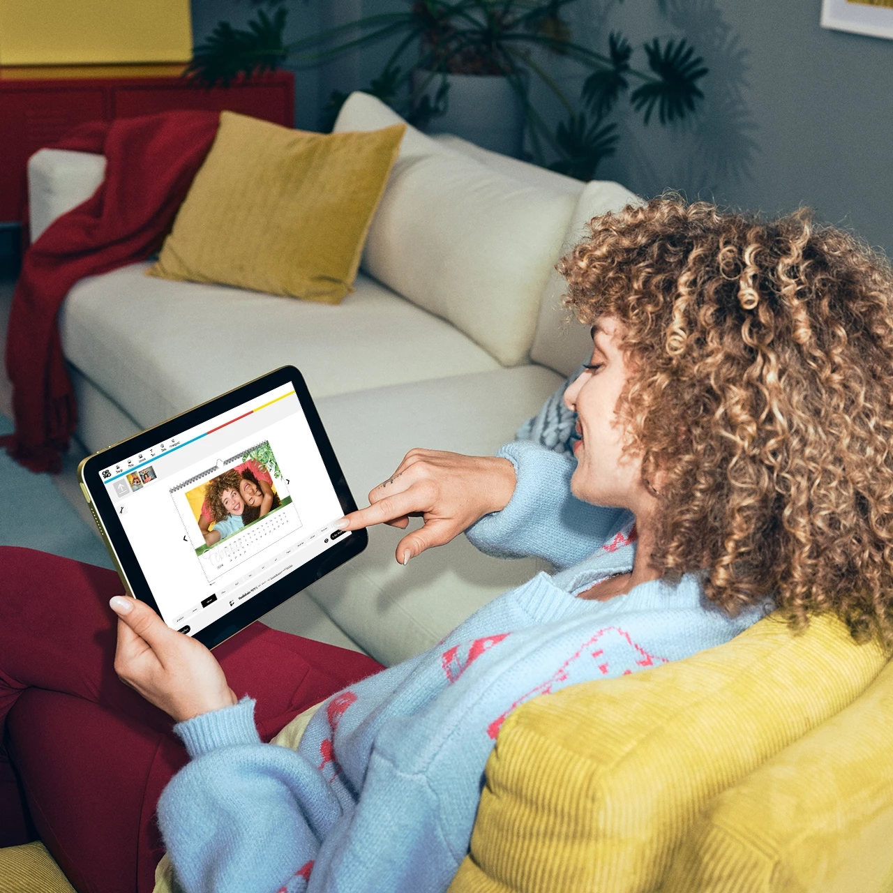 Eine lockige Frau gestaltet einen Kalender im ORWO-Fotodesigner von zuhause aus, auf der Couch sitzend, mit einem Tablet und sieht dabei tiefenentspannt aus.