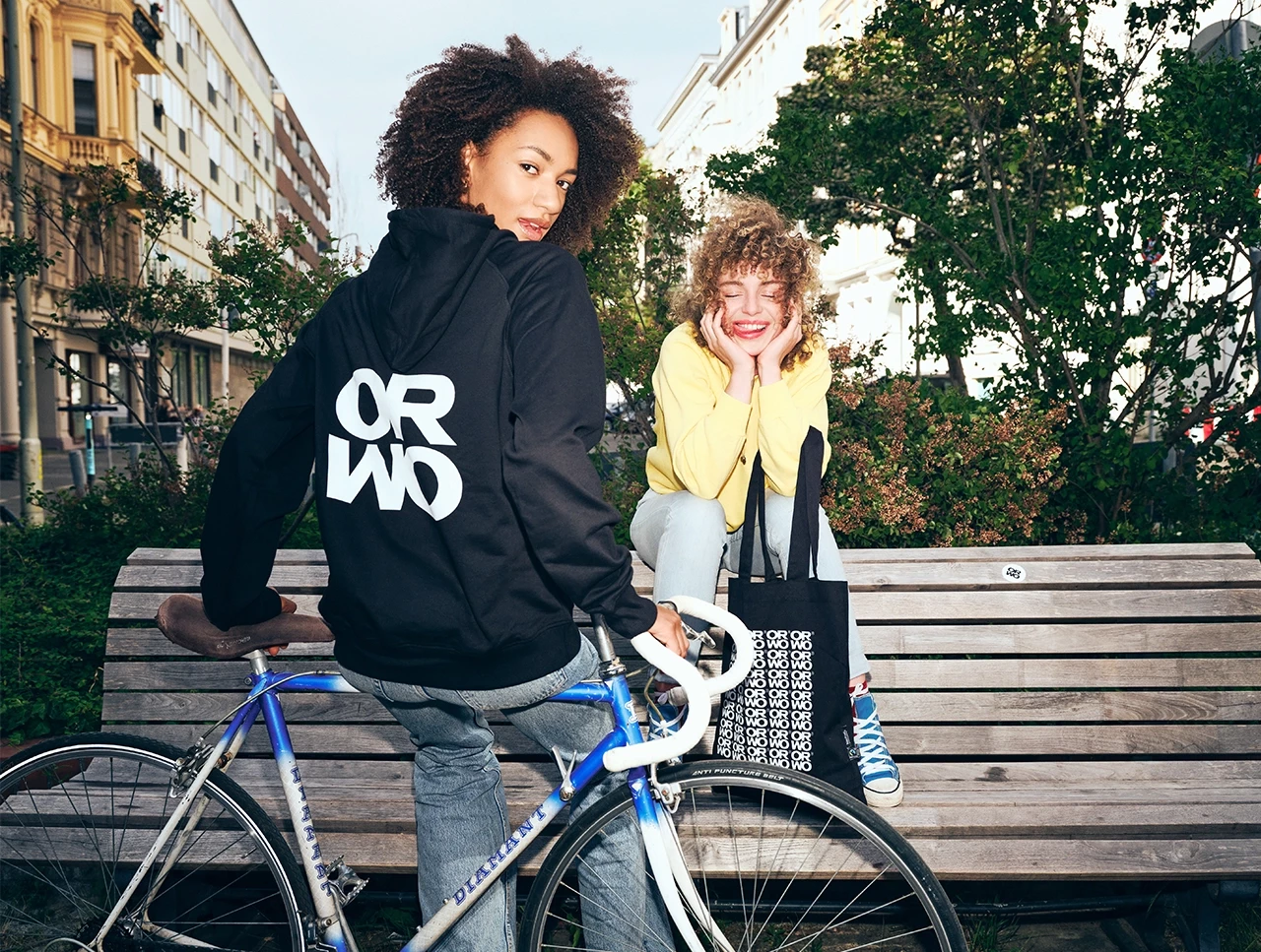 Zwei Frauen im Park: Eine sitzt lässig auf einer Banklehne mit ORWO-Stofftasche, die andere steht mit Fahrrad im Vordergrund und trägt einen ORWO-Hoodie.