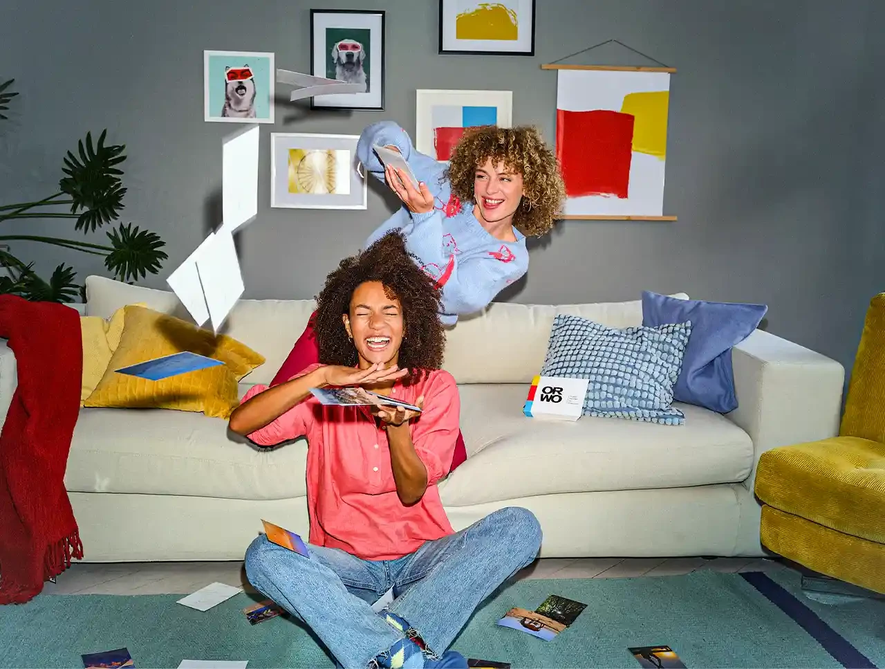Fotos, hochwertiges Fotopapier, UV-beständig, scharfe Bilder Zwei Frauen lassen Fotos regnen und haben sichtlich Spaß dabei. Die Bilder wurden bei ORWO bestellt. Auf der Couch liegt das stylische Verpackungsmaterial.