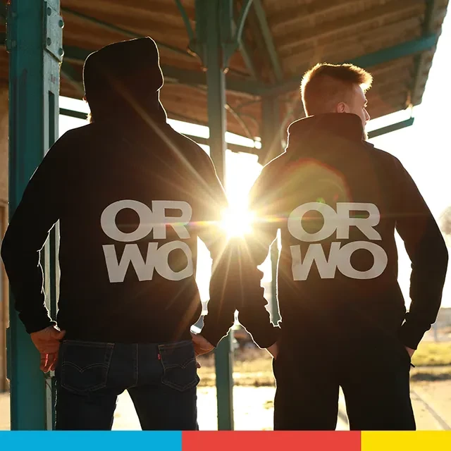 Sweatjacke mit großem ORWO-Logo auf der Rückenseite Ein Gegenlichtfoto, auf dem zwei Herren mit dem Rücken zur Kamera stehen. Sie präsentieren das große ORWO-Logo auf dem Rücken der Sweatjacke mit epischem Lense Flare.