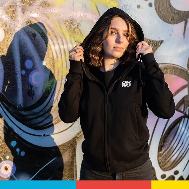 Sweatjacke mit ORWO-Print, doppellagige Kapuze, hochwertig Zu sehen ist eine Frau, die vor einer mit Sprühkunst verzierten Wand steht und die stylische ORWO Sweatjacke in Szene setzt. Bestelle jetzt dein ORWO-Piece.