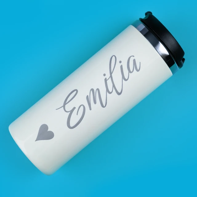 Weißer Thermobecher mit graviertem Schriftzug „Emilia“ und Herzsymbol auf hellblauem Hintergrund.