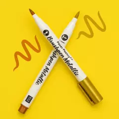 Zwei metallische Brushpens in Gold und Bronze auf gelbem Hintergrund, mit gemalten Farbschlieren in Metallic-Tönen.