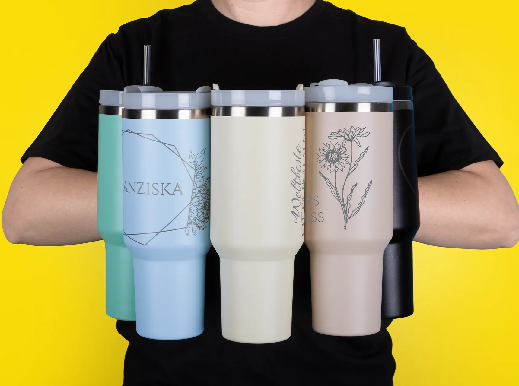 Fünf große Thermobecher in Stanley Cup Optik, die vor dem Körper gehalten werden, mit Henkel und Strohhalm. In fünf Farben erhältlich.