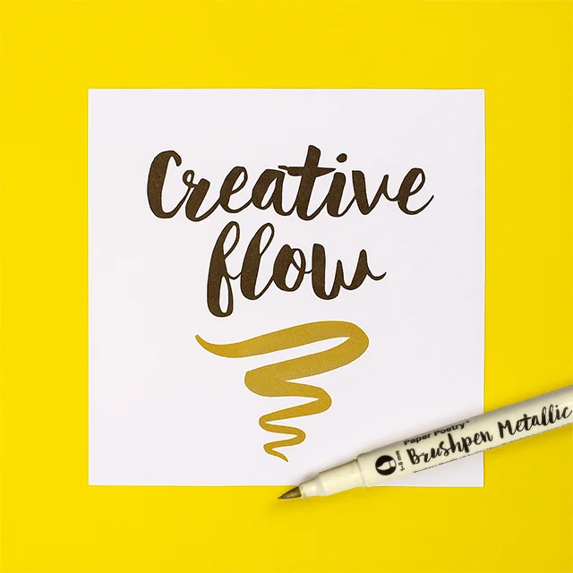 Goldener Brushpen auf gelbem Hintergrund, daneben eine Karte mit der Aufschrift „Creative Flow“ in glänzender Metallic-Schrift.