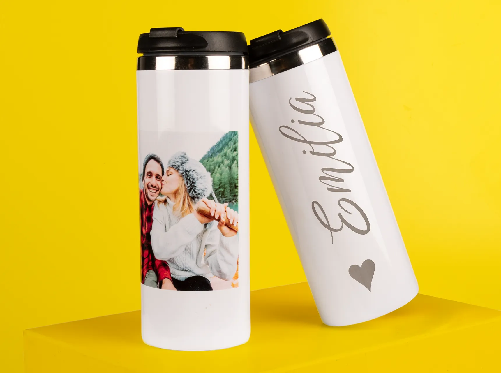 Zwei weiße Thermobecher auf gelbem Hintergrund – einer mit Foto eines Paares, der andere mit dem gravierten Schriftzug „Emilia“ und Herzsymbol.