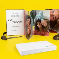 Foto Leporello, stehend, mit Geschenkverpackung Ein Leporello steht auf einer gelben Fläche. Es ist mit Fotos von Freundinnen personalisiert. Im Vordergrund liegt die weiße Geschenkverpackung.