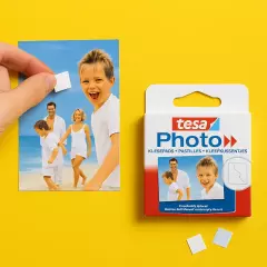 Hand befestigt ein Foto mit einem tesa Photo Klebepad, daneben die Verpackung auf gelbem Hintergrund.